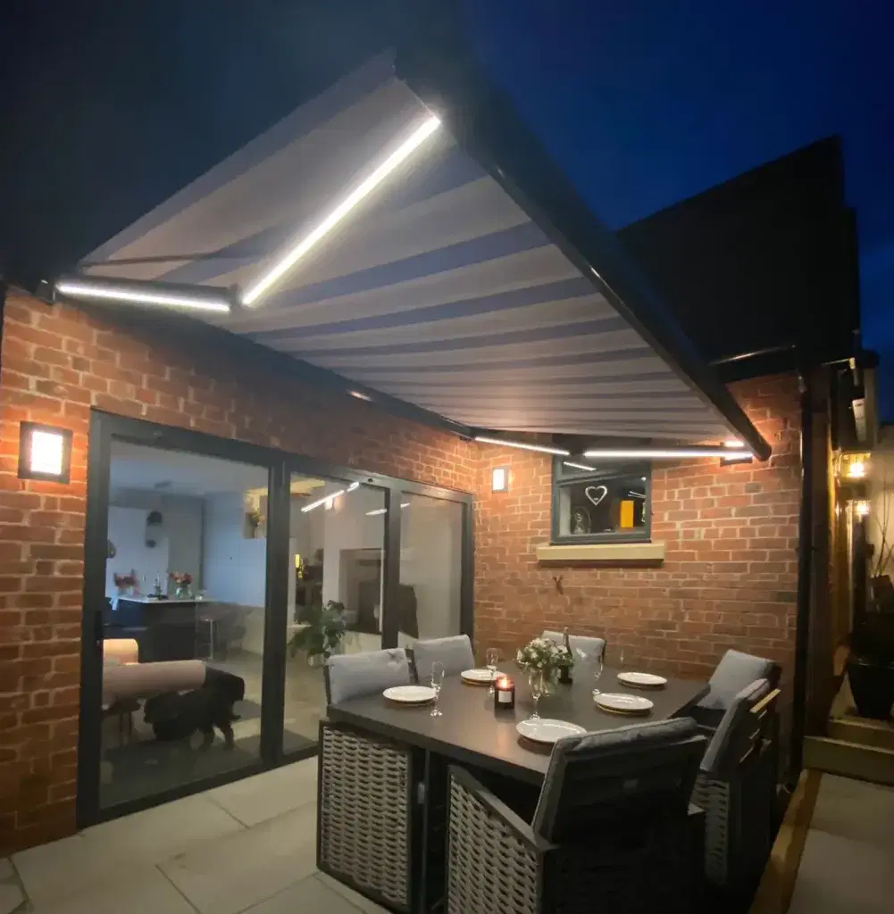 A stripy awning covering a patio with tables and chairs at night with built in lights
